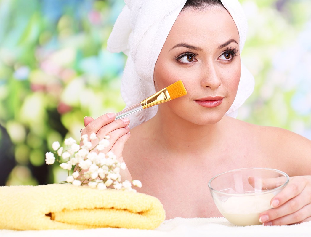 Beautiful young woman with cream for face mask towel on her head on bright background